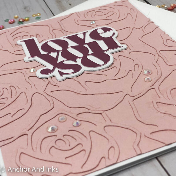 pink valentine card with die cut roses background and pink metallic wording that says love you so